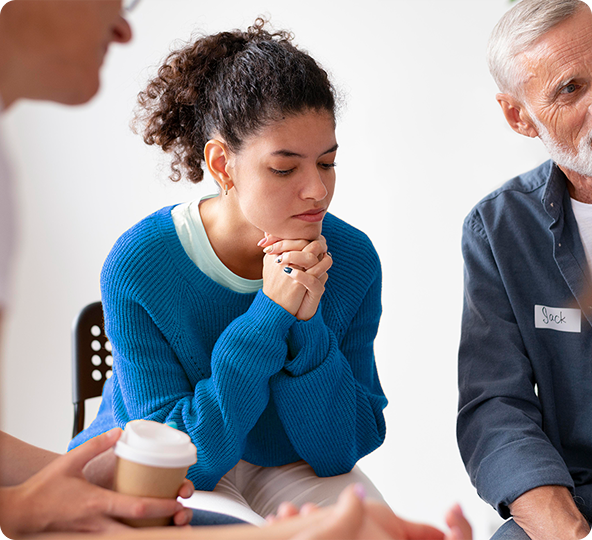 Woman in blue sweater in a support group, hands clasped, listening.