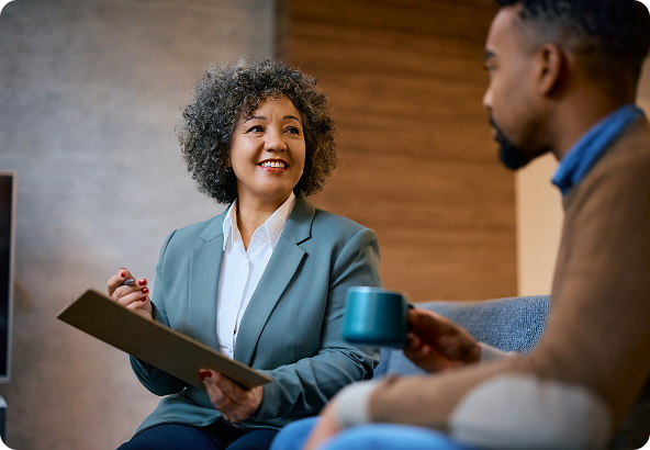 Woman in a blazer smiles and holds a clipboard, talking to a person holding a mug in a cozy setting.