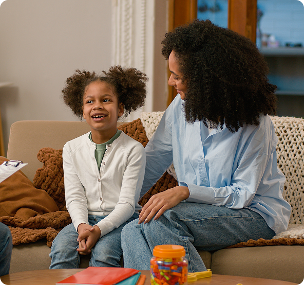 Woman smiles at a child sitting together on a couch. The child has curly hair and wears a white sweater.