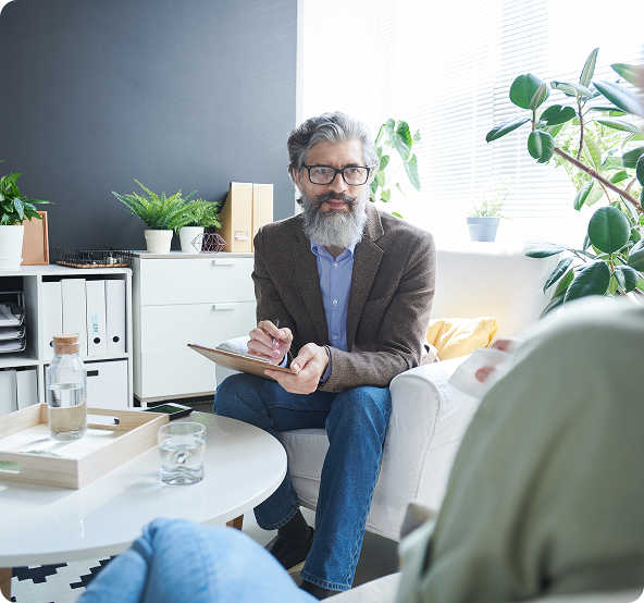 Therapist with a clipboard, speaking to a seated client in a bright office.