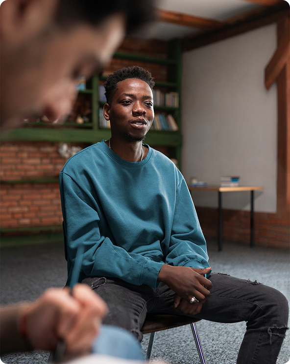 Man in teal sweatshirt speaking, sitting in chair. Another person's shoulder and arm in foreground. Room with bookcase.