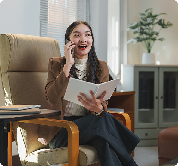 Woman on phone, smiling, holding notebook, sitting in office chair.