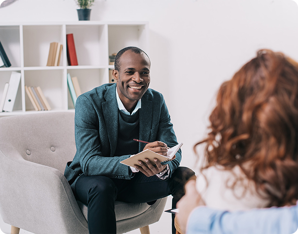 Therapist smiling, seated, talking to patient in an office. Bookshelf in the background.