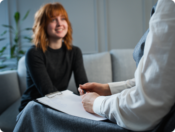 Woman smiling during therapy session; therapist holds clipboard, setting is a living room.