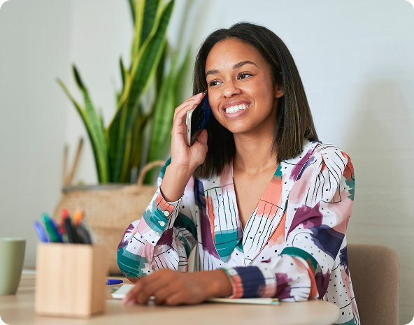 Woman smiling while talking on a phone at a desk with pens, a plant, and a notebook.