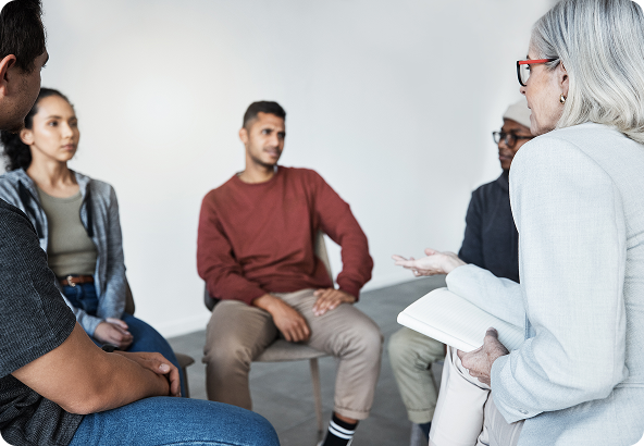 Group therapy session: People seated in a circle listening to a facilitator holding a notebook.
