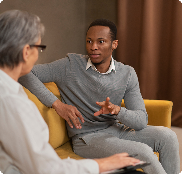Man in gray sweater talking, seated on yellow couch, to person with glasses.