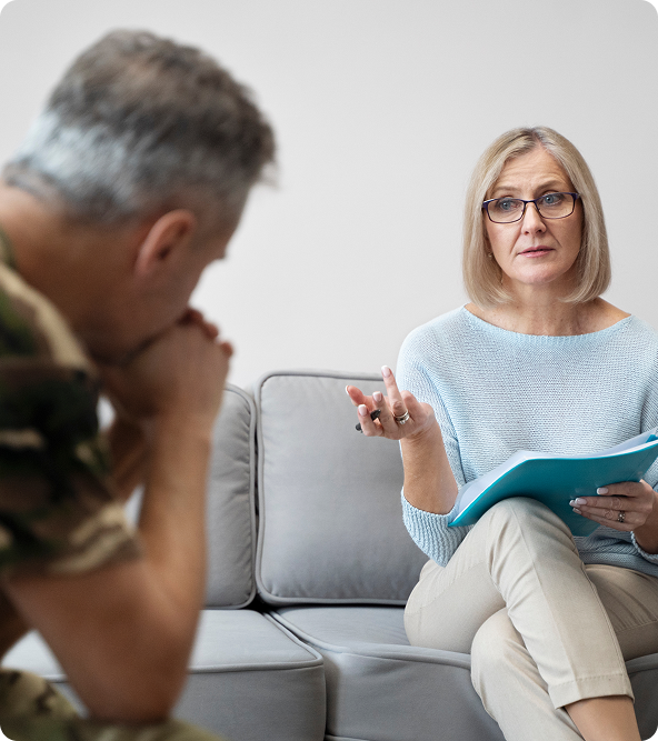 Therapist with notepad gesturing, talking to a person in military uniform on a couch.