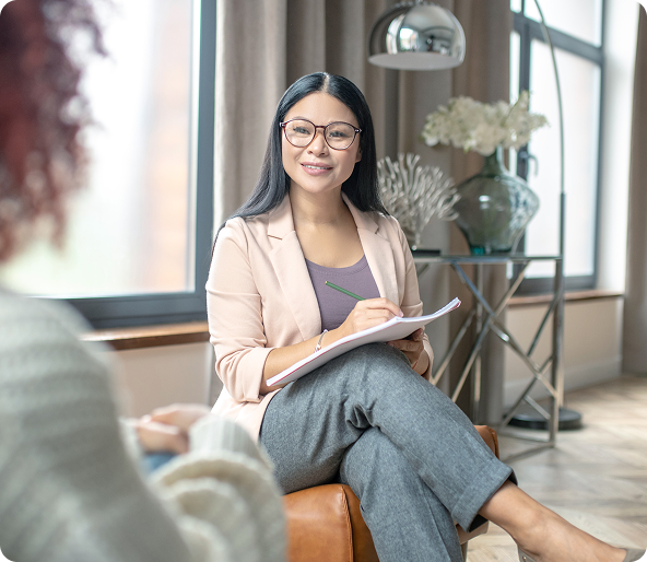 Therapist in glasses, seated, smiles, takes notes. Client blurred, indoor setting.