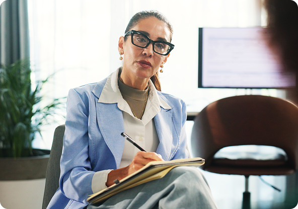 Woman in glasses, writing in notebook, appears to be listening intently. Interior office setting, light blue jacket.