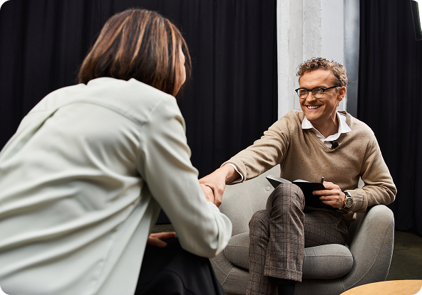 Man in glasses shakes hands with person, sitting in armchairs. Smiling, light background.