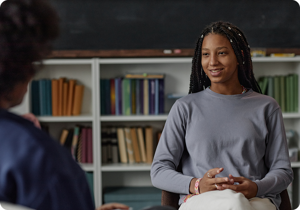 Woman in gray shirt smiles while talking to another person in a therapy session; bookshelves in background.