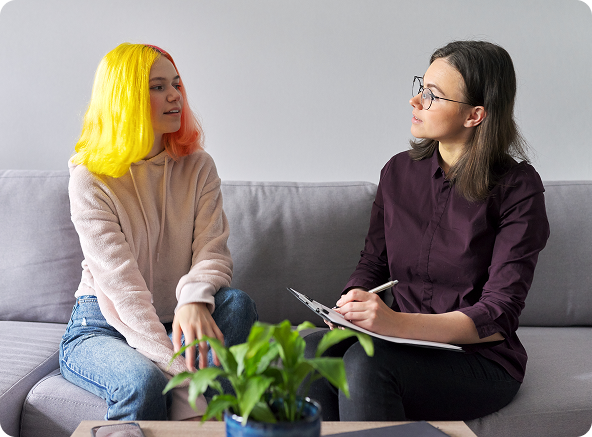 A person with colorful hair speaks to a person holding a notepad, seated on a couch indoors.