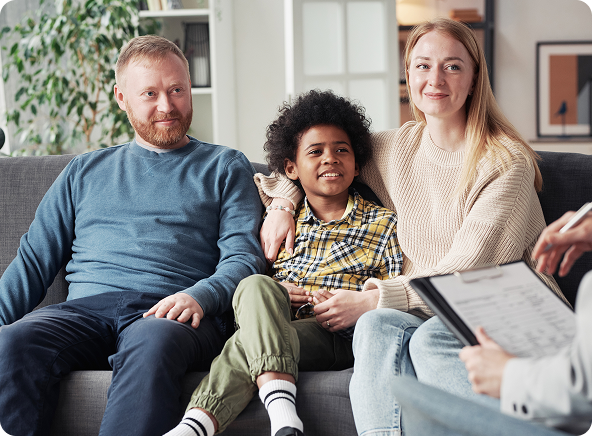 Family of three on a couch during a therapy session, smiling at the person holding a clipboard.