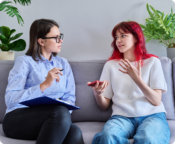 Woman with glasses and a woman with red hair sit on a couch, one speaking and the other taking notes.