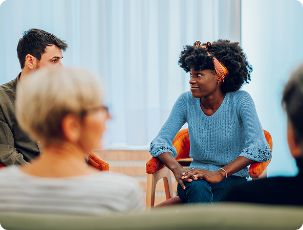 People sitting in a circle, engaged in a group discussion. A woman in a blue sweater appears to be speaking.