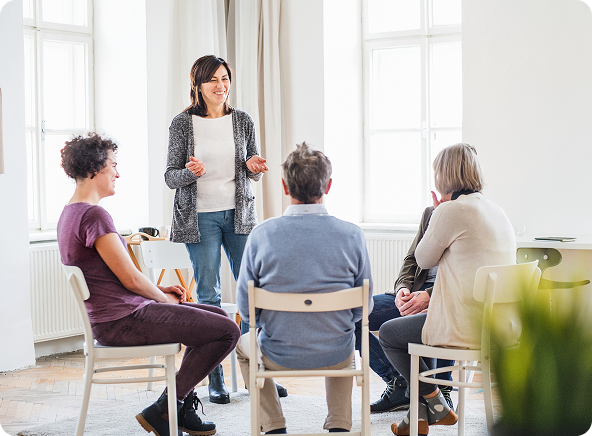 Group therapy session: Woman holding paper addresses seated people in a circle. Bright room, natural light.