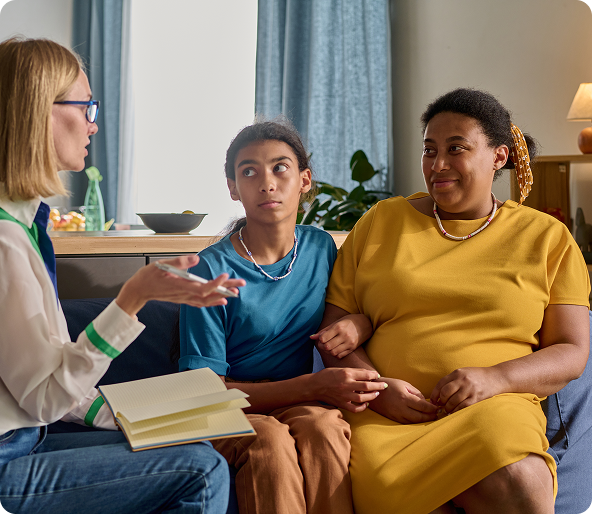A woman with glasses talks to a mother and a teenager on a couch.