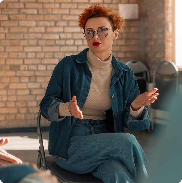 Woman in glasses gesturing, speaking in a room with a brick wall.
