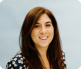 Woman with long brown hair smiles at the camera, wearing a dark floral print top, blue background.