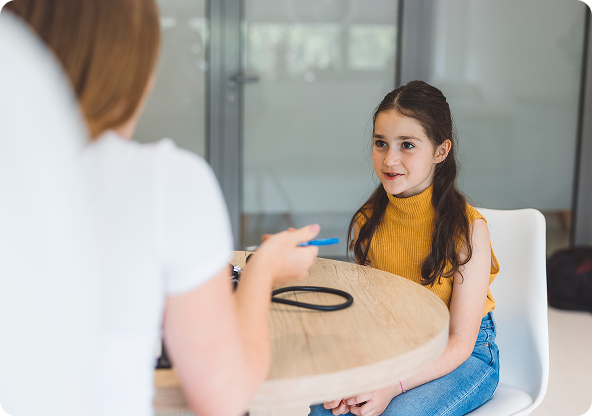 Woman speaking to a girl seated at a table. Girl smiles, wearing yellow top and jeans. Modern interior.