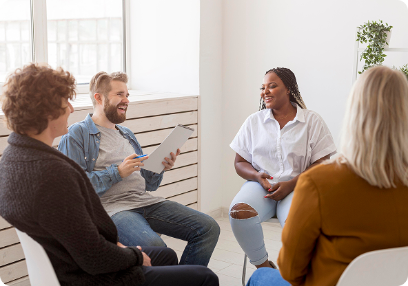 Four people seated in a circle, engaged in conversation. Light-filled room, white chairs.