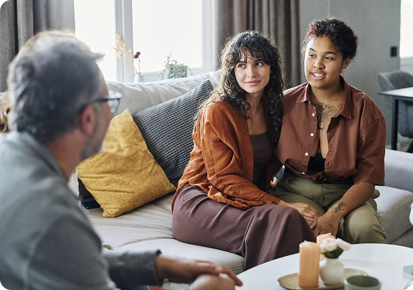 Two women sit on a sofa, looking at a man, in a living room.