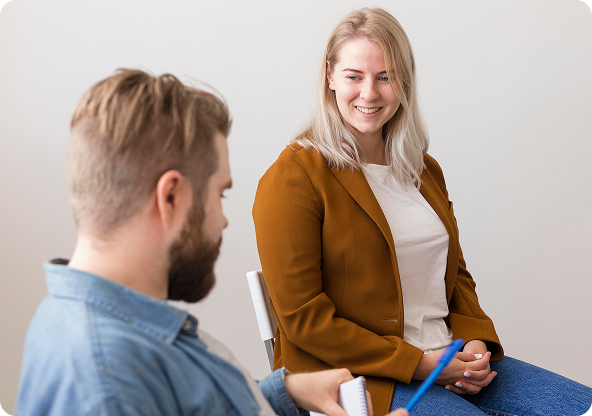 Woman in brown blazer smiles while speaking to a man in blue shirt holding a notepad.