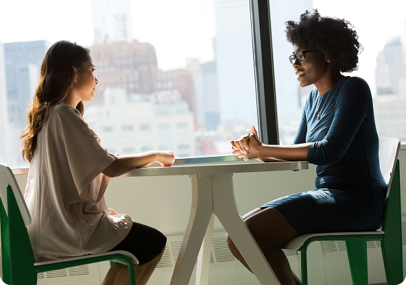 Two people seated at a table by a window, engaged in conversation. Sunlight fills the room.