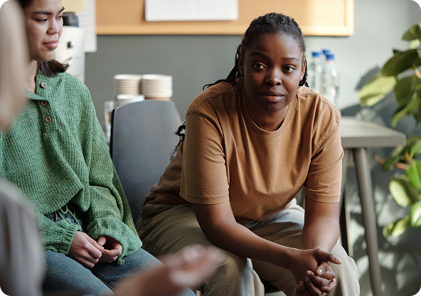 Woman in tan shirt at a group therapy session; looks at someone. Another person in green nearby.