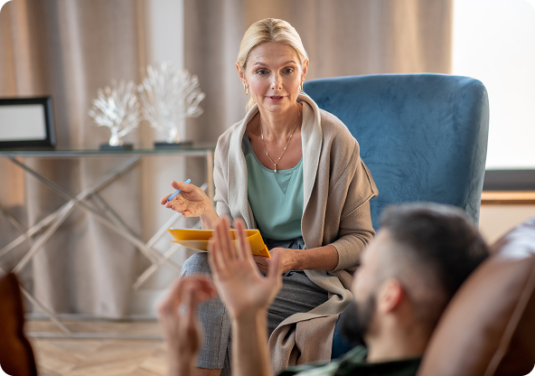 Therapist in light cardigan gestures, talking to patient reclining in armchair. Neutral setting.