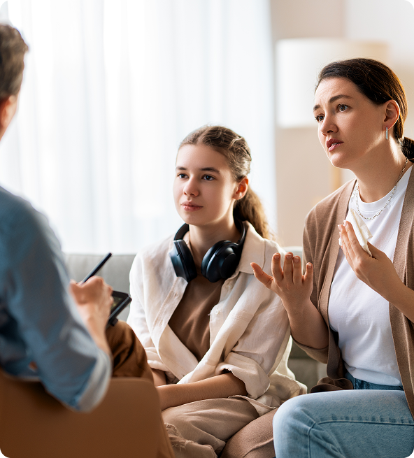 Therapist counseling a mother and teenage daughter; mother gestures, daughter listens with headphones.