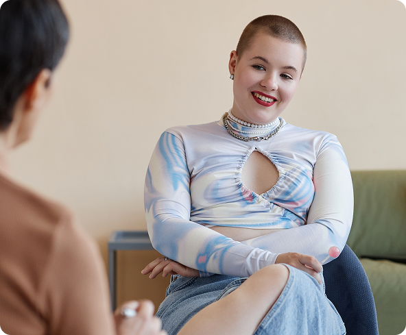 Woman with shaved head smiles, sits cross-legged, wearing patterned top, talking to another person.