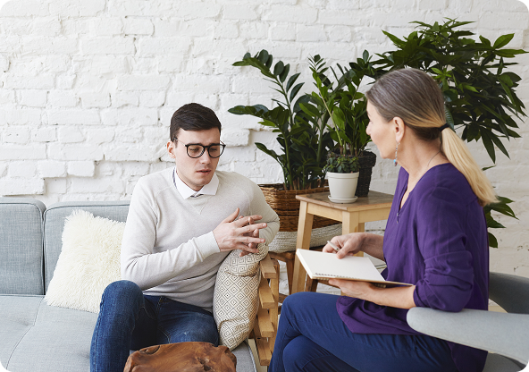 A person in glasses talking to another person with a notebook, seated on a couch in a room with plants.