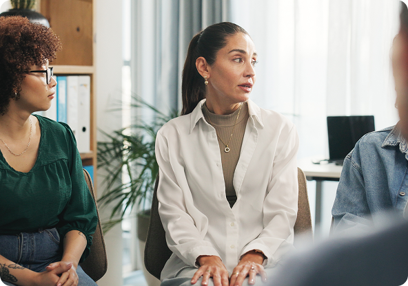 People in a group therapy session, one woman speaking, others listening, in a bright room.