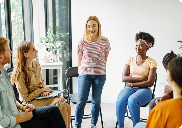 Group therapy session: woman speaking, surrounded by seated listeners in a bright room.