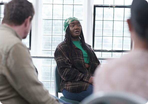 Woman in a group setting, speaking. Wearing a green crop top, plaid shirt, and headscarf, seated, windows in background.