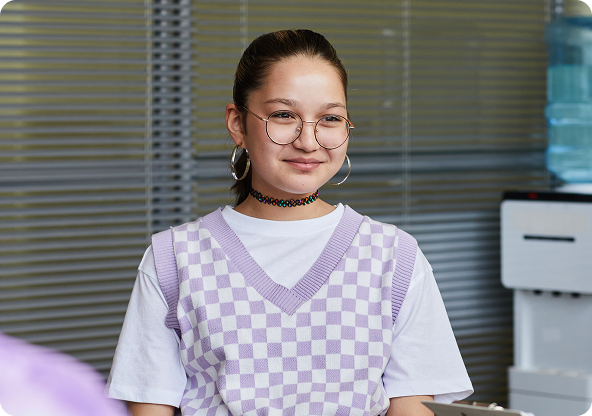 Woman in glasses and choker smiles, wearing a purple checkered vest and white shirt. Beside a water cooler.