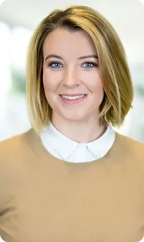 Woman with blonde hair smiling, wearing a tan sweater over a white collared shirt.
