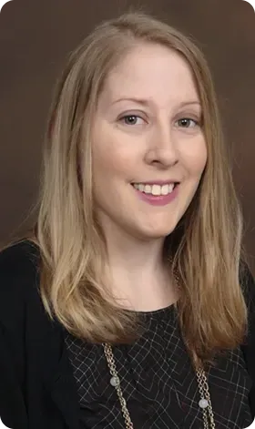 Blond woman with a slight smile, wearing a black top with a patterned inset, against a brown backdrop.