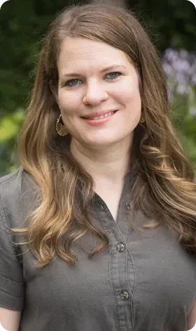 Woman with long, wavy brown hair smiles while wearing a green button-down shirt. Outdoors with blurred background.