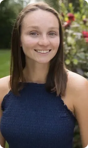 Woman smiling, wearing a blue halter top, standing outdoors with foliage in the background.