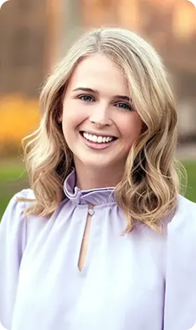 Woman with blonde wavy hair, smiling, wearing a light purple blouse outdoors.