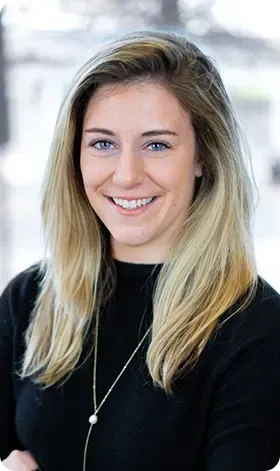 Woman with blonde hair, smiling, wearing a black top and a necklace, looking at the camera.