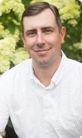 Man in white shirt smiling outdoors in front of leafy green background.