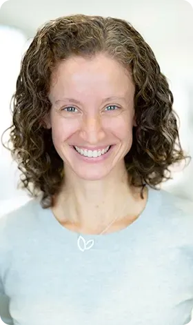 Woman with curly hair smiles broadly, wearing a light blue shirt and necklace.