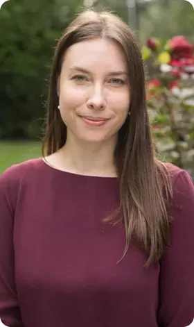 Woman with long brown hair, wearing a burgundy top, smiling outdoors.