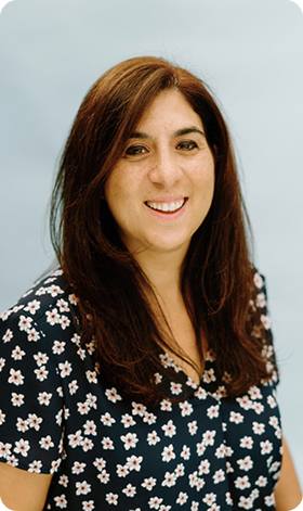Woman smiling, with long brown hair, wearing a dark blue floral patterned top, against a light blue background.