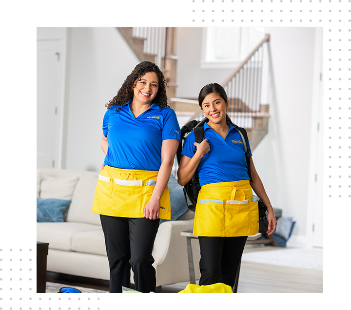 Two cleaning staff in blue uniforms and yellow aprons stand smiling in a bright living room.