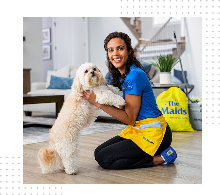 Woman in blue shirt and yellow apron kneels with a fluffy white dog in a bright living room